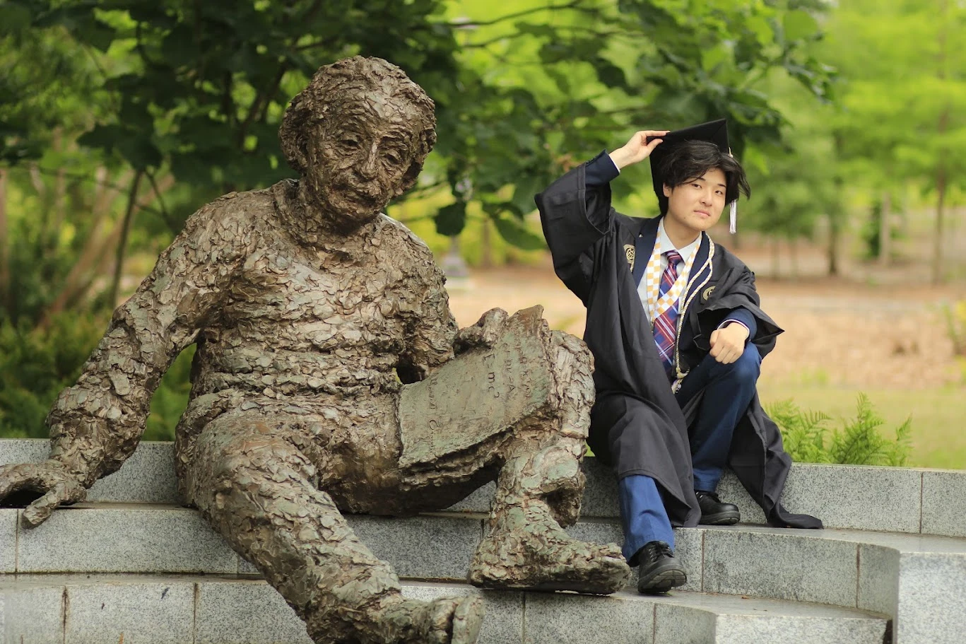 Justin Park in graduation attire seated beside a statue outdoors